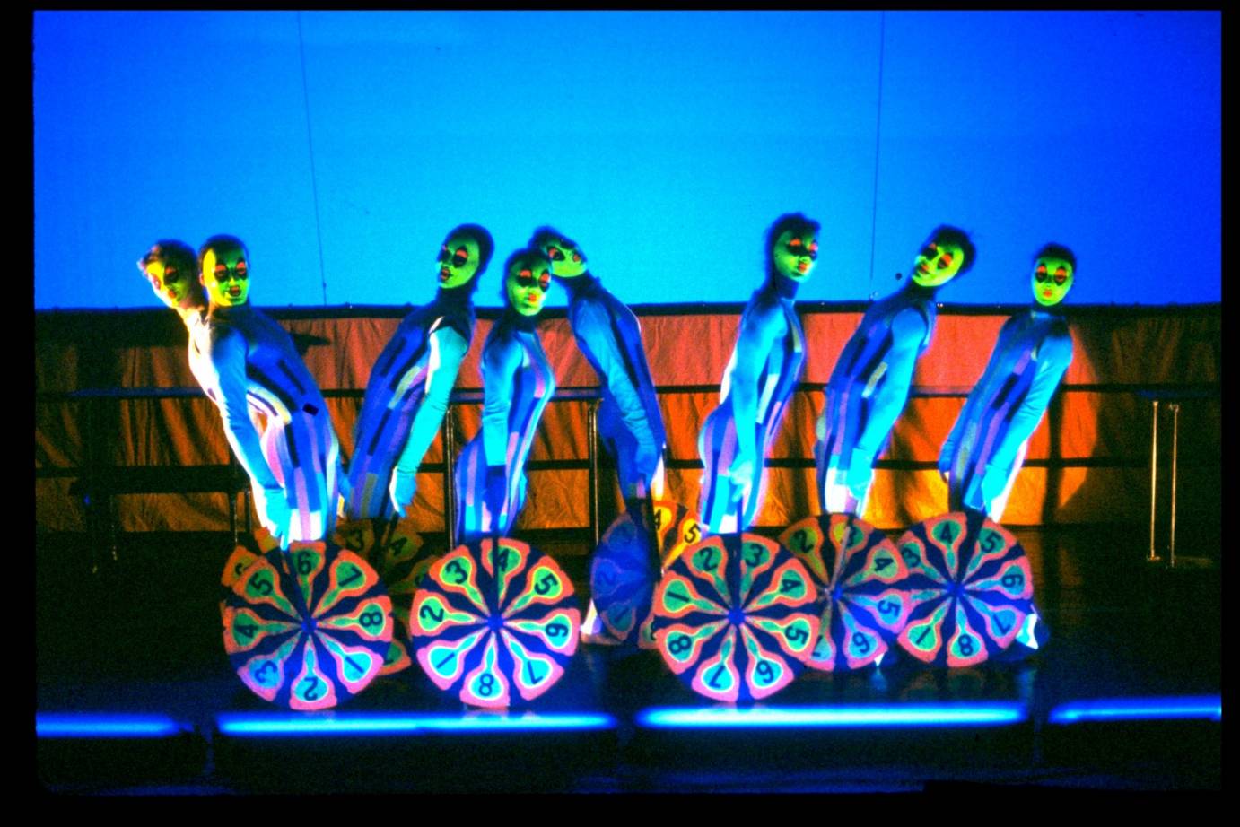 Dancers resembling clowns with neon green faces and standing on or behind pin wheel-looking installations.